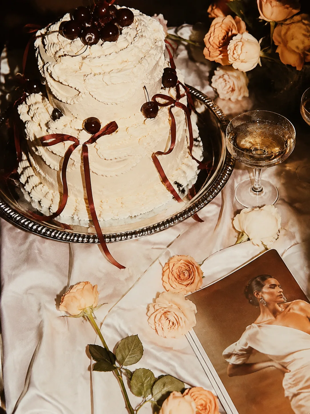A cherry-topped wedding cake beside roses and a coupe of champagne.