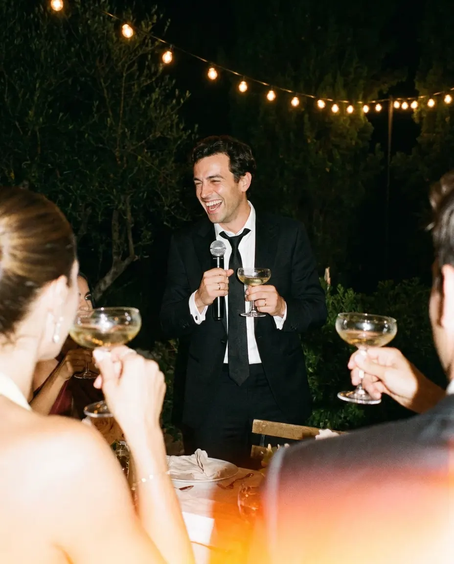 A best man laughing mid-toast with a microphone and a glass of champagne, filmed from a guest’s table.
