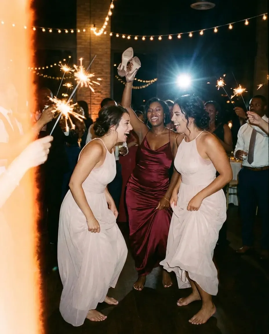 Three bridesmaids dancing barefoot among sparklers, one waving her heels overhead.