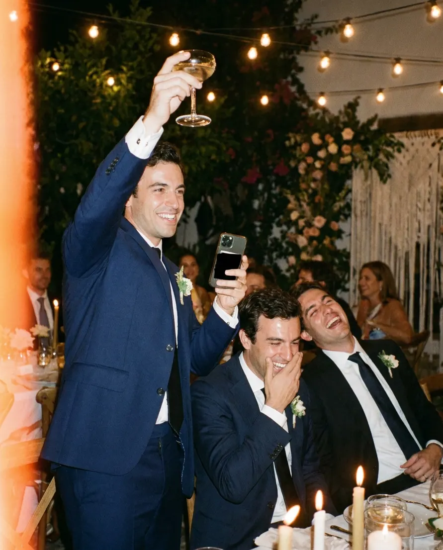 The best man raising a glass mid-toast, phone with his notes in hand, the groomsmen cracking up.